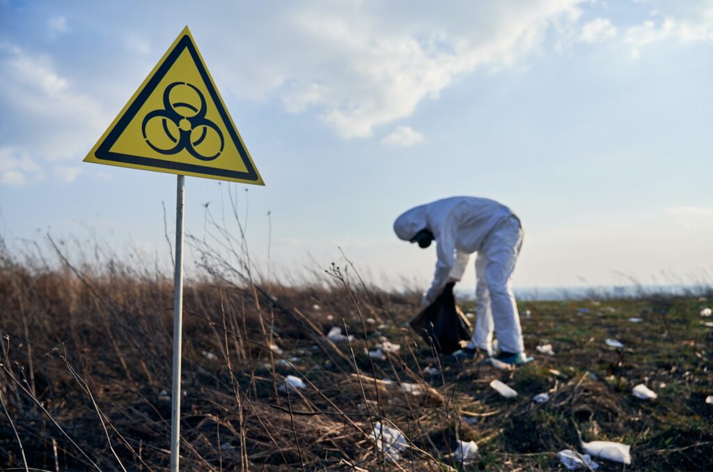Researcher in protective suit collecting plastic garbage into black waste bag outdoors on sunny day